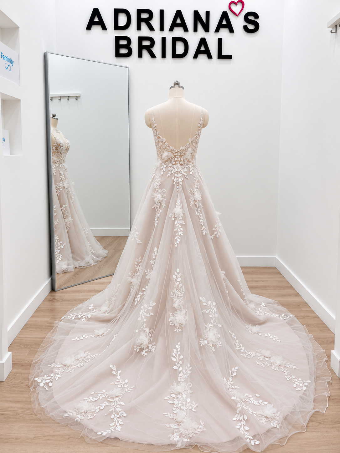 A bridal gown displayed on a mannequin in a boutique fitting room, with a long train spread across a wood floor and a large mirror showing the back of the dress. The dress is a soft blush-beige tulle with white floral lace applique.