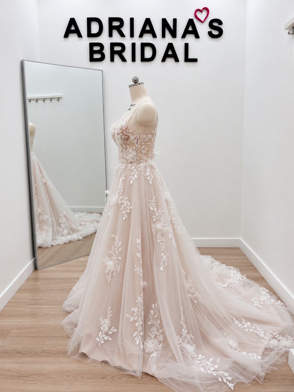 A pale blush pink A-line wedding gown with white floral lace applique, displayed on a dress form in a bridal shop. A large floor mirror reflects the back of the gown, and the text 'ADRIANA'S BRIDAL' is visible on the wall above.