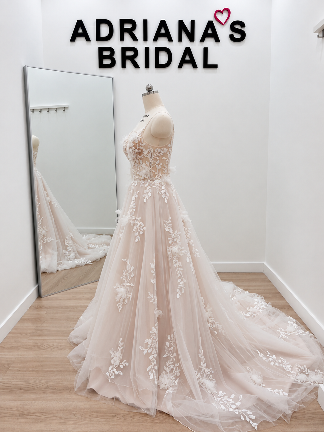 A pale blush pink A-line wedding gown with white floral lace applique, displayed on a dress form in a bridal shop. A large floor mirror reflects the back of the gown, and the text 'ADRIANA'S BRIDAL' is visible on the wall above.